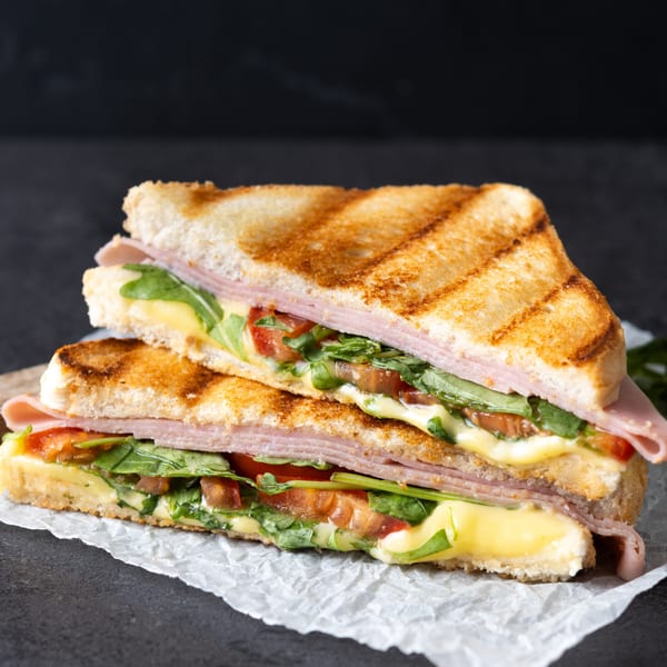 Two halves of a grilled Goodi's sandwich with ham, melted cheese, arugula, and tomatoes.