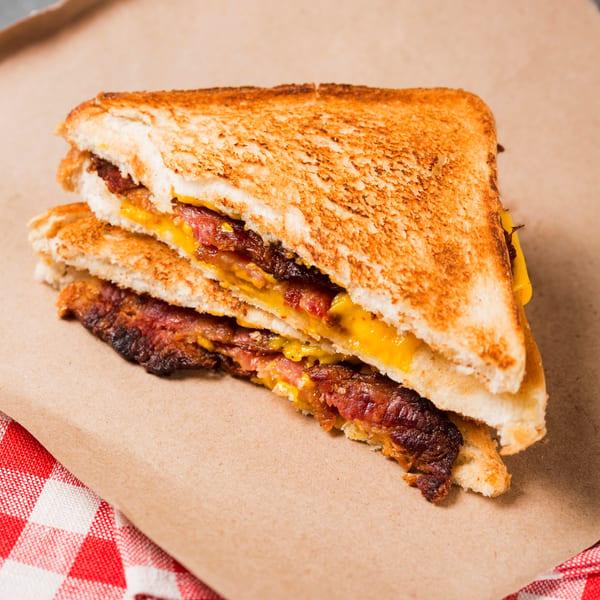 Toasted sandwich with melted cheese and crispy bacon, served on brown paper with a red and white checkered napkin.