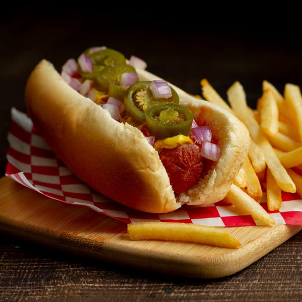 A hot dog with jalapeños, red onions, and mustard, served with french fries on a wooden board.