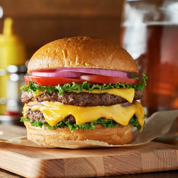A double cheeseburger with lettuce, tomato, and red onion on a wooden board with a drink.